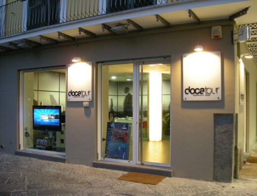 Vetrine di DOCATOUR, agenzia di viaggi specializzata in tour in moto e accessori per motociclisti, con insegna luminosa e monitor.