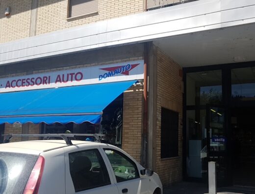 Domauto negozio di ricambi e accessori auto con tenda blu, situato in un edificio di mattoni, con un'auto bianca parcheggiata di fronte.