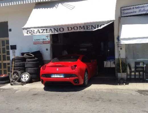 Immagine dell'officina di Domenico Graziano con una Ferrari rossa parcheggiata di fronte. Si notano pneumatici impilati e l'insegna "GRAZIANO DOMENIC". Servizi di riparazione e pneumatici Kumho offerti.