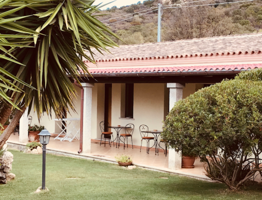 Veranda esterna di una casa in Sardegna, con tavolini e sedie, circondata da un giardino curato e vegetazione mediterranea. Ideale per rilassarsi all'aperto.