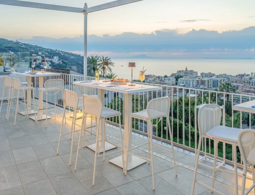 Terrazza panoramica con tavoli alti e sedie bianche, vista sul mare e sulla città al tramonto. Arredamento moderno e curato, ideale per aperitivi e cene all'aperto.