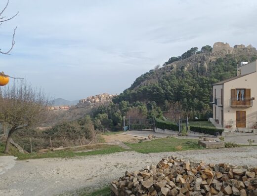 Paesaggio panoramico presso Donna Vì Hotel, con vista sul castello medievale in cima alla collina, case del paese, albero di cachi con frutti maturi e legna da ardere in primo piano.