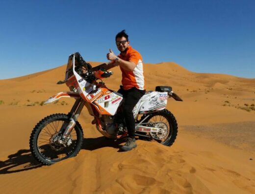 Un motociclista sorridente su una moto da rally KTM nel deserto del Sahara, con dune di sabbia e cielo azzurro. Moto accessoriata per viaggi avventura e competizioni. Servizi e marketplace dottormoto.it