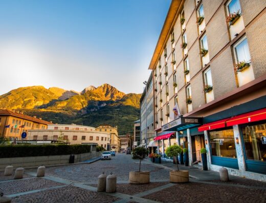 Paesaggio urbano ad Aosta con l'Hotel Duca D'Aosta in primo piano, montagne sullo sfondo illuminate dal sole al tramonto e pavimentazione in pietra.