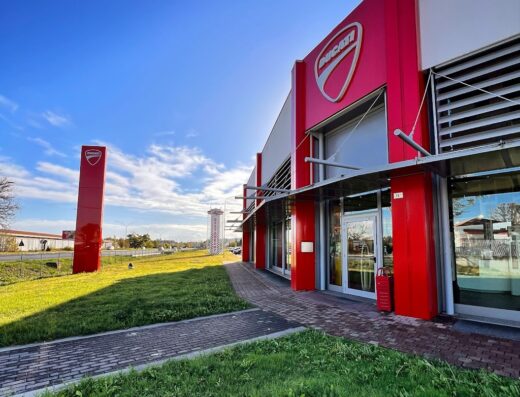 Esterno del Ducati Factory Store, con insegna rossa e logo Ducati ben visibili, prato verde e cielo azzurro. Accessori e abbigliamento moto disponibili.
