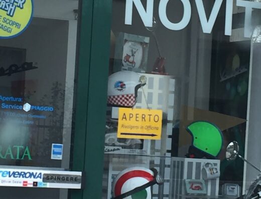 Vetrina di Due Ruote Verona Srl con esposizione di caschi e accessori per moto Vespa, cartello "Aperto", e indicazioni per il servizio Piaggio. Novità e offerte per scooter.