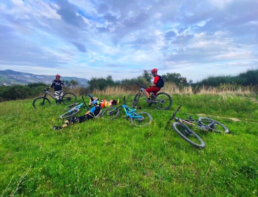Gruppo di ciclisti in pausa su un prato verde a DueruoteForli, con mountain bike appoggiate e due ciclisti sdraiati sull'erba, cielo nuvoloso sullo sfondo