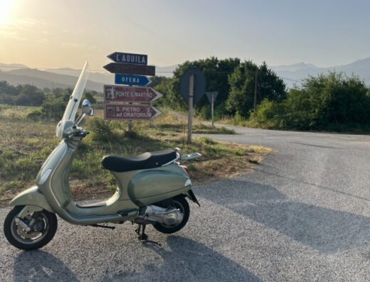 Noleggio scooter in Abruzzo: Vespa verde parcheggiata su strada con cartello indicatore per L'Aquila, Ofena, Ponte S. Martino e S. Pietro ad Oratorium. Ideale per tour e gite.