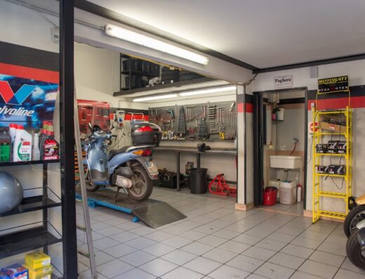 Interno di un'officina moto Eddy Motor Bike: scooter blu su rampa di sollevamento, attrezzi a parete, scaffali con olio Valvoline e batterie Motobatt, lavandino.