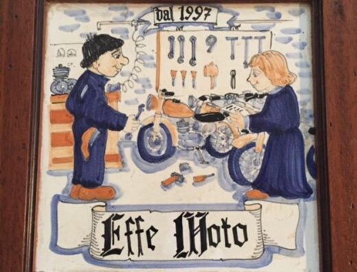 Disegno su ceramica di Effe Moto dal 1997, raffigurante due meccanici che lavorano su una moto d'epoca, con attrezzi appesi sullo sfondo.