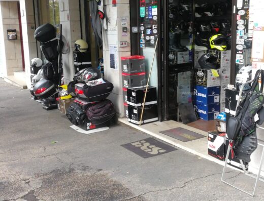 Negozio di accessori per motociclisti Eliomoto a Roma, con caschi, abbigliamento tecnico e valigie laterali esposti all'esterno. Vetrina con adesivi e prodotti Premier.