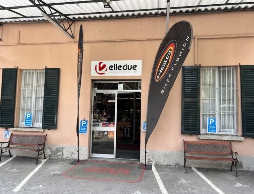 Ingresso del negozio Elledue, rivenditore di accessori e abbigliamento per motociclisti, con insegna e bandiere promozionali di marchi di settore.