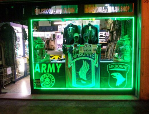 Vetrina illuminata di Ello Militaria a Maistrello Roberta & co., con esposizione di abbigliamento e accessori militari, inclusi simboli "Army" e "Airborne", evocando un'atmosfera da negozio specializzato per appassionati.
