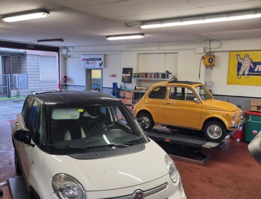 Interno di un'autofficina Emmeci Gommista con una Fiat 500L bianca in primo piano e una Fiat 500 gialla su un ponte sollevatore.