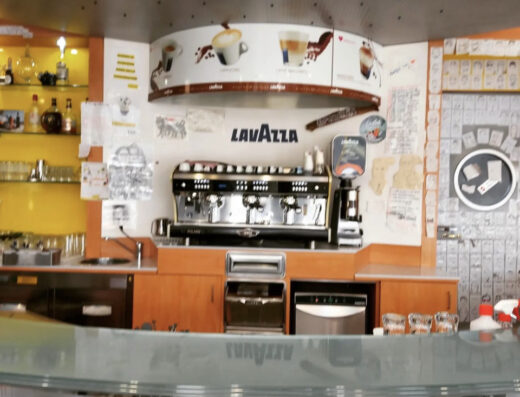 Interno dell'Eni Café & Shop: bancone bar con macchina da caffè Lavazza professionale, scaffali con liquori e decorazioni murali.