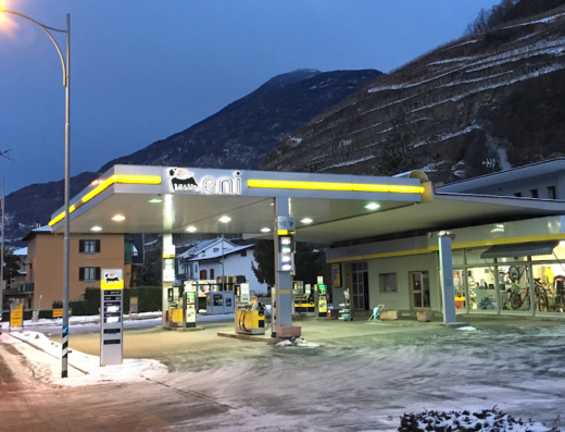 Eni Station Cioccarelli in Sondrio, Italy, with a view of the mountains in the background. The station offers fuel and possibly a motorcycle repair or accessory shop, visible through the windows.
