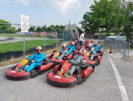 Gruppo di piloti pronti alla partenza in pista di kart a Entrata Pista Verde, con caschi e tute da corsa, per un'esperienza di guida emozionante e sicura.