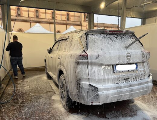 Un SUV Nissan X-Trail bianco è ricoperto di schiuma in un autolavaggio Errelle CarWash in Via Appia, mentre un uomo lo lava.