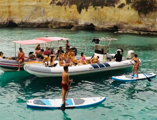 Gita in barca e stand-up paddleboarding al mare: persone si rilassano su gommoni e fanno SUP vicino alla costa rocciosa durante un servizio di escursioni e noleggio barche.