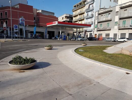 Esso Barletta: stazione di servizio con motocicletta parcheggiata, auto in fila e architettura urbana circostante. Servizi di rifornimento e accessori per viaggi.