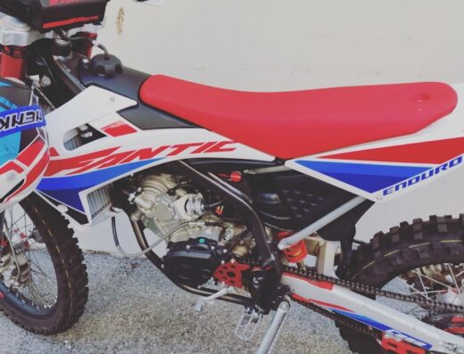 Moto da enduro Fantic bianca, rossa e blu con casco da cross appoggiato, ideale per il motocross e il tempo libero, disponibile su Eurobike Srl.