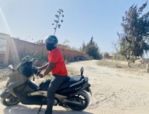 Un uomo su uno scooter TMAX nero in un paesaggio assolato. Indossa un casco e una maglietta rossa. Il percorso sembra essere in una zona rurale o costiera.