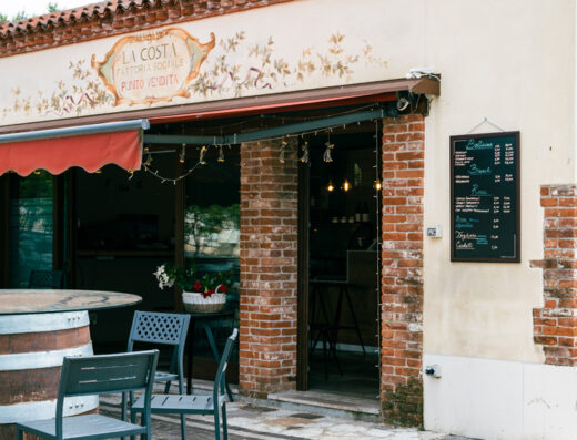 Fattoria Sociale La Costa exterior view with rustic facade, outdoor seating, featuring tables made from barrels, and a menu board displaying local dishes.