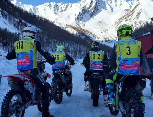 Piloti di motocross della Federazione Motociclistica Italiana CoRe Piemonte pronti per la gara sulla neve, con montagne innevate sullo sfondo.
