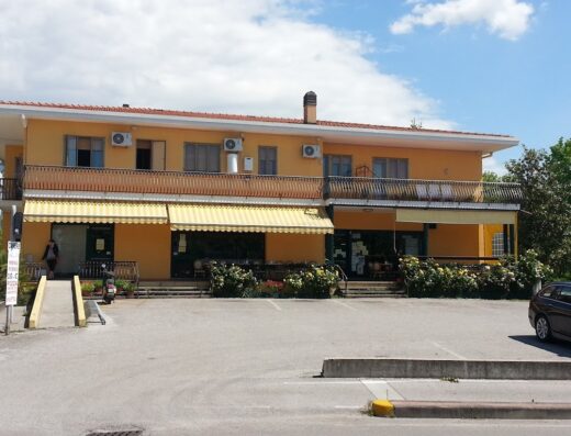 Edificio giallo a due piani con balcone, tende da sole e parcheggio antistante. Presenza di un cartello menù e di un piccolo scooter parcheggiato di fronte.