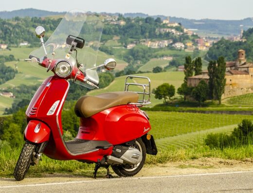 Tour in Vespa nelle Langhe: una Vespa rossa con parabrezza parcheggiata su una strada di campagna con vista panoramica sulle colline e sui vigneti del Piemonte.