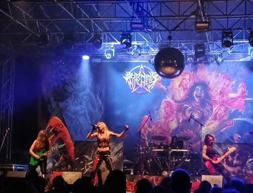 Concerto live della band heavy metal Burning Witches con musiciste sul palco e pubblico che assiste all'esibizione musicale.