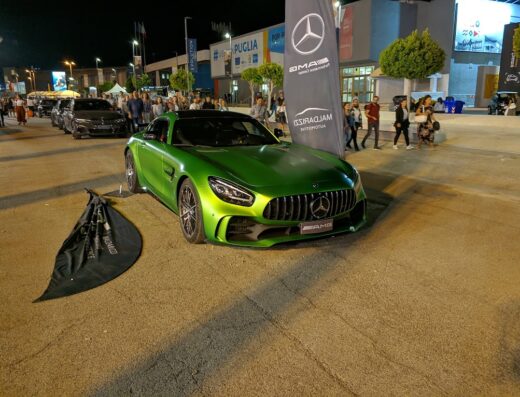 Fiera del Levante: una lussuosa Mercedes-AMG verde metallizzato esposta all'ingresso orientale, con visitatori sullo sfondo e un banner del concessionario Malaguzzi Automotive.