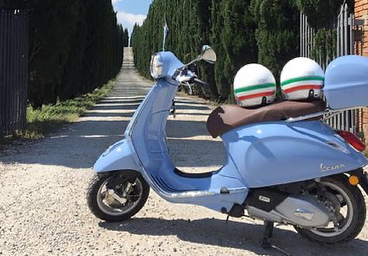 Noleggio Vespa a Firenze: Scooter azzurro parcheggiato su strada sterrata con due caschi tricolore e bauletto, ideale per esplorare la Toscana.