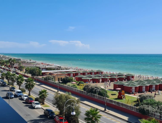Vista panoramica del Fly Decó Hotel, con la spiaggia affollata, il mare turchese e le cabine colorate. Traffico sulla strada e palme.