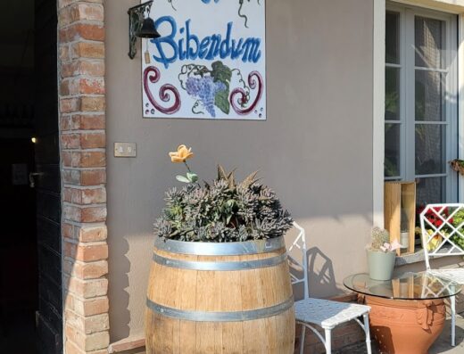 Esterno di Cascina Sciulun di Franco Conterno con un cane che riposa accanto a una botte di legno usata come fioriera, sotto l'insegna "Nunc est bibendum".