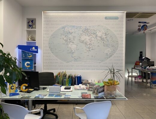 Ufficio di Frida Viaggi con scrivania ordinata, computer, logo Costa Crociere e grande mappa del mondo con scritte "Buon Viaggio" in diverse lingue.
