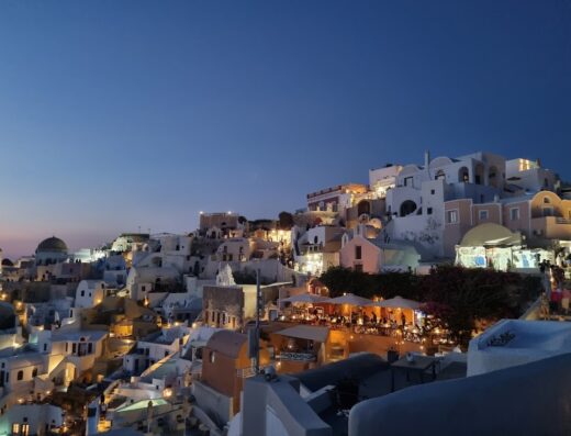 Veduta notturna di Santorini, Grecia, con le sue iconiche case bianche illuminate, ristoranti con tavoli all'aperto e cielo crepuscolare, perfetta per viaggi e vacanze.