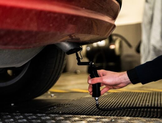 A mechanic is testing the emissions of a red car at Garage Delfino. The exhaust pipe is connected to a handheld emissions testing device.