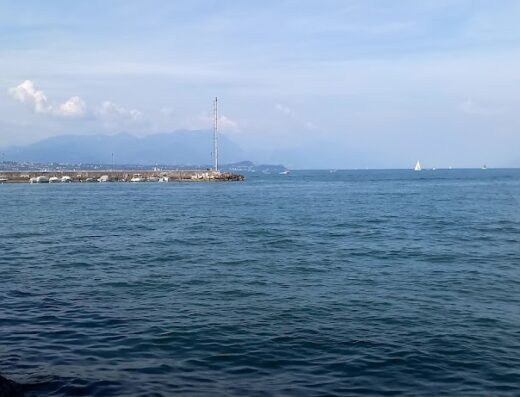 Noleggio moto d'acqua a Desenzano del Garda: Vista panoramica del lago con molo, barche ormeggiate e montagne sullo sfondo. Servizi di affitto e noleggio disponibili.