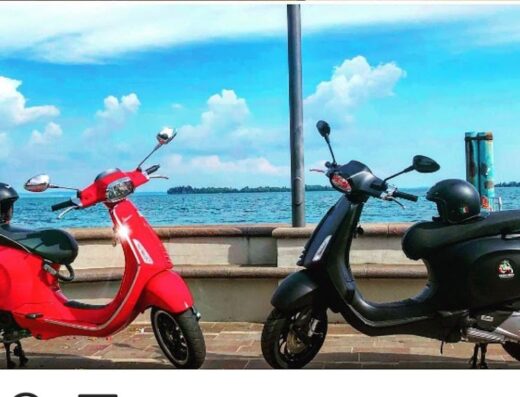 Noleggio Vespa sul Lago di Garda: Due scooter Vespa, uno rosso e uno nero, parcheggiati con caschi, pronti per esplorare il lago.