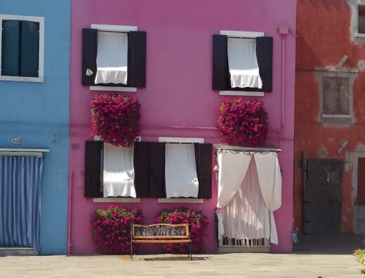 Colorata casa a Burano, Italia, con facciata rosa, finestre con persiane marroni, fiori rossi e una panchina in legno, tipica architettura veneziana.