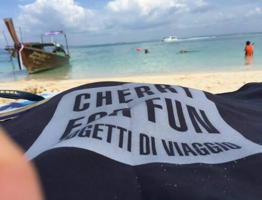 Spiaggia tropicale con barca tradizionale thailandese, mare cristallino e telo mare con scritta "Cherry for Fun, Oggetti di Viaggio". Accessori viaggio disponibili da Gattinoni Travel Store Vignola.