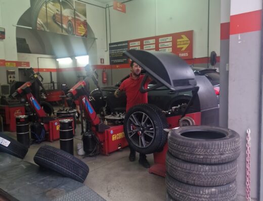 Gommista al lavoro nell'officina Giannini F.Lli S.R.L., impegnato nella sostituzione pneumatici con attrezzatura professionale. Gomme nuove e usate visibili.