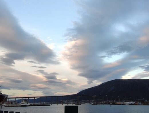 Panorama di Tromsø, Norvegia, con una nave nel porto, un ponte sullo stretto e montagne sullo sfondo sotto un cielo nuvoloso al tramonto. Ideale per tour in moto e viaggi.