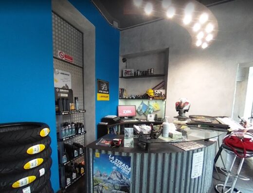 Interno del negozio Gieffe Moto: banco reception con logo SYM, scaffale con pneumatici e accessori moto, espositori di lubrificanti e ricambi, illuminazione moderna.