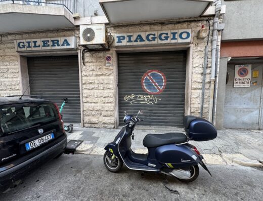 Foto di una concessionaria Gilera Piaggio con saracinesche chiuse, una Vespa blu parcheggiata davanti e un'auto nera di lato.
