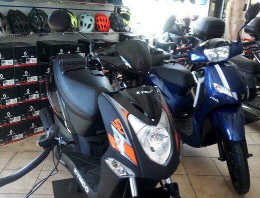 Primo piano di scooter Kymco nero e blu in un negozio di accessori moto, con caschi da motociclista e accessori Gipiemme sullo sfondo, per vendita e assistenza.