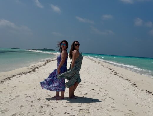 Due donne in abiti estivi posano su una spiaggia di sabbia bianca alle Maldive, con acque turchesi che si estendono all'orizzonte sotto un cielo azzurro nuvoloso; viaggio e vacanze con GIRAMONDO VIAGGI SCANDIANO