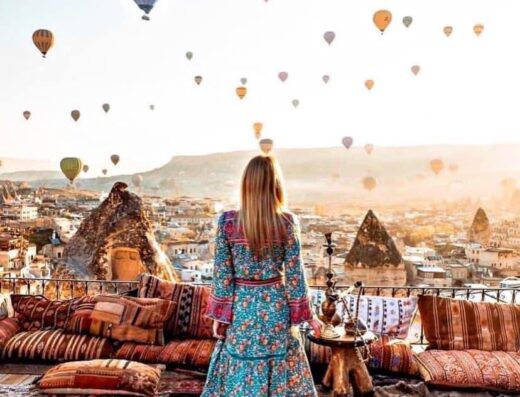 Una donna in abito lungo ammira il panorama di Cappadocia con mongolfiere, seduta su cuscini e tappeti. Girotour Sardegna, agenzia viaggi Cagliari.