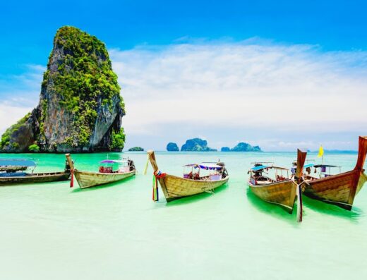 Barche tradizionali thailandesi longtail ormeggiate in acque turchesi con un isola rocciosa sullo sfondo, meta turistica popolare in Asia.